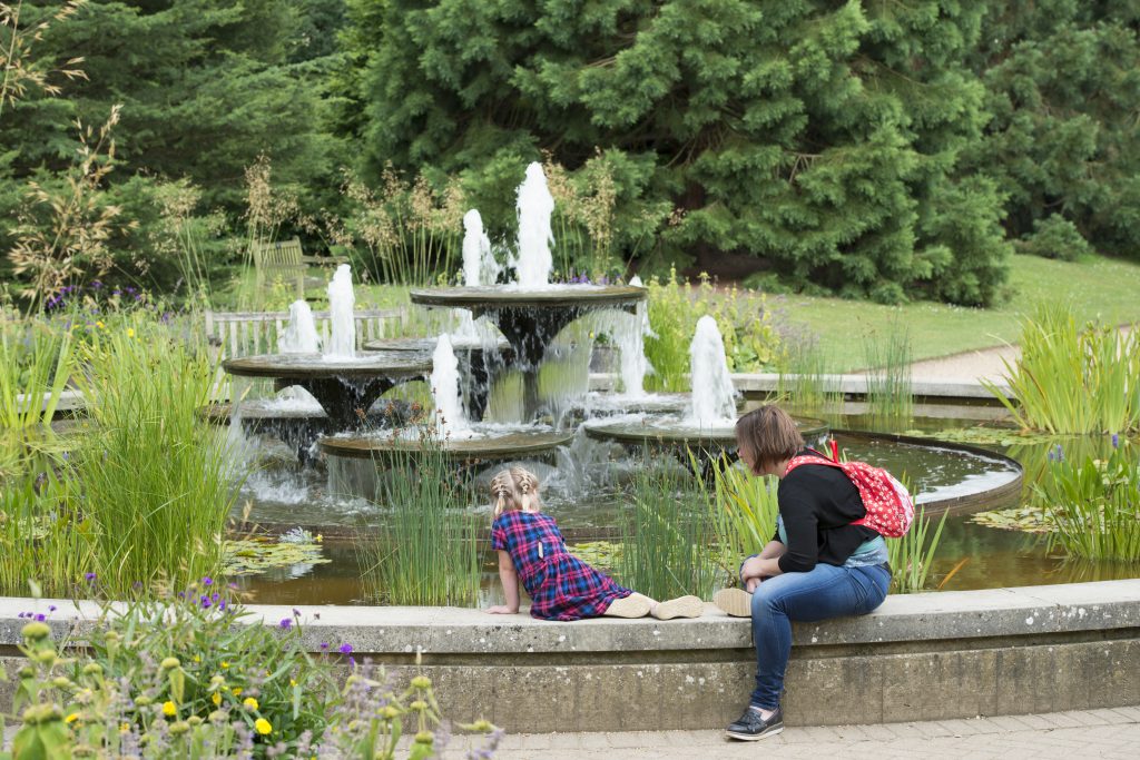 A woman and child sit on the edge of the David Mellor fountain, looking in to the water.