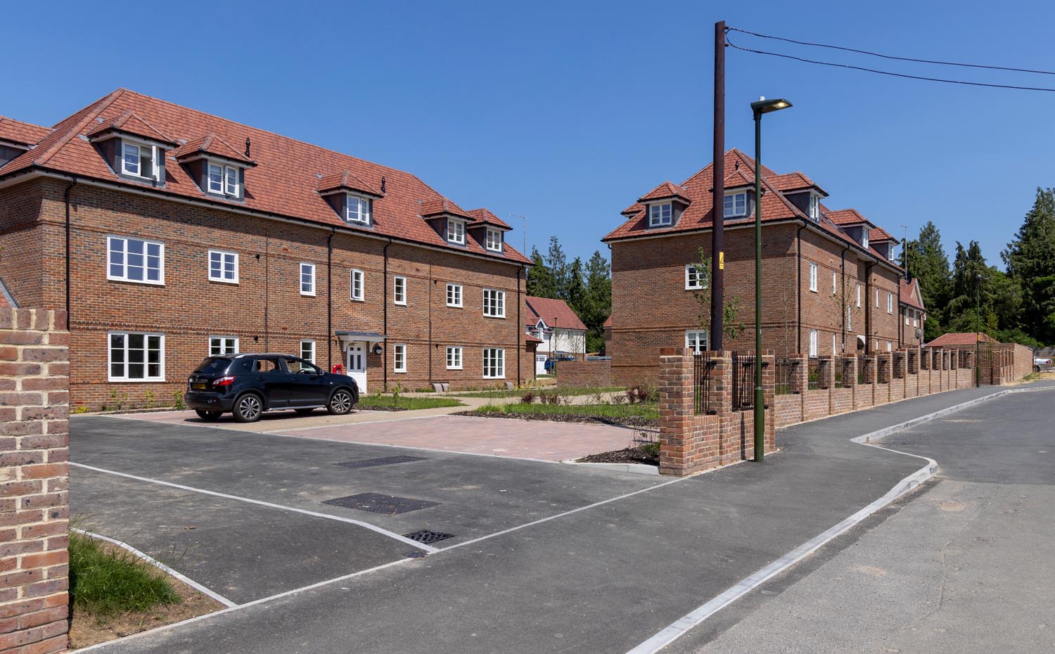 Residential street scene showing modern brick apartment buildings with pitched tiled roofs, a marked parking area, and paved access under clear daylight.