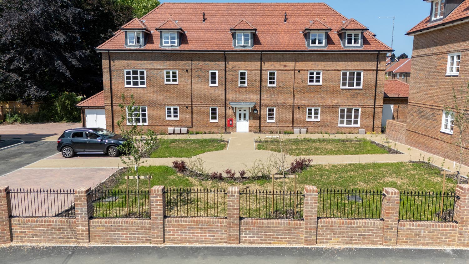 Front view of modern brick residential buildings positioned behind a landscaped communal green with pathways, planted borders, and low metal fencing