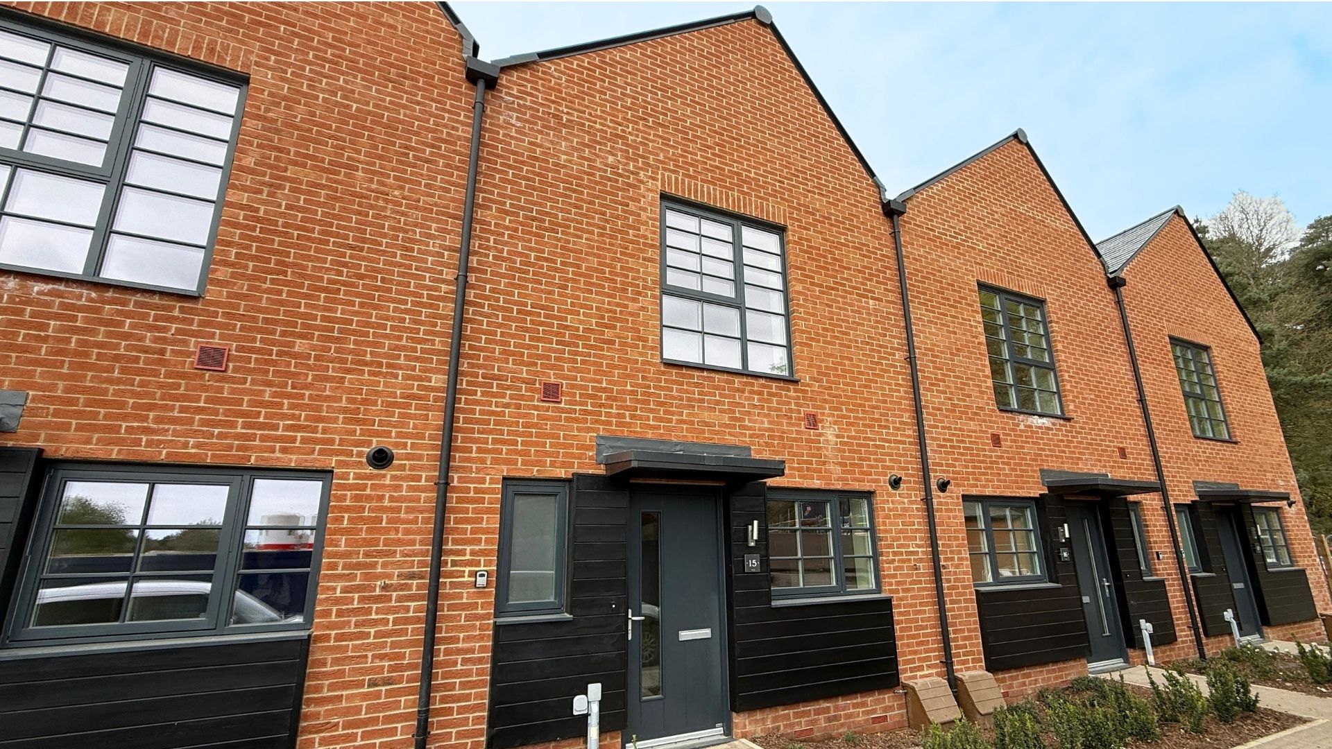 Modern brick house with a black front door, pitched roof, white‑framed windows, and minimal front planting along a paved residential frontage.