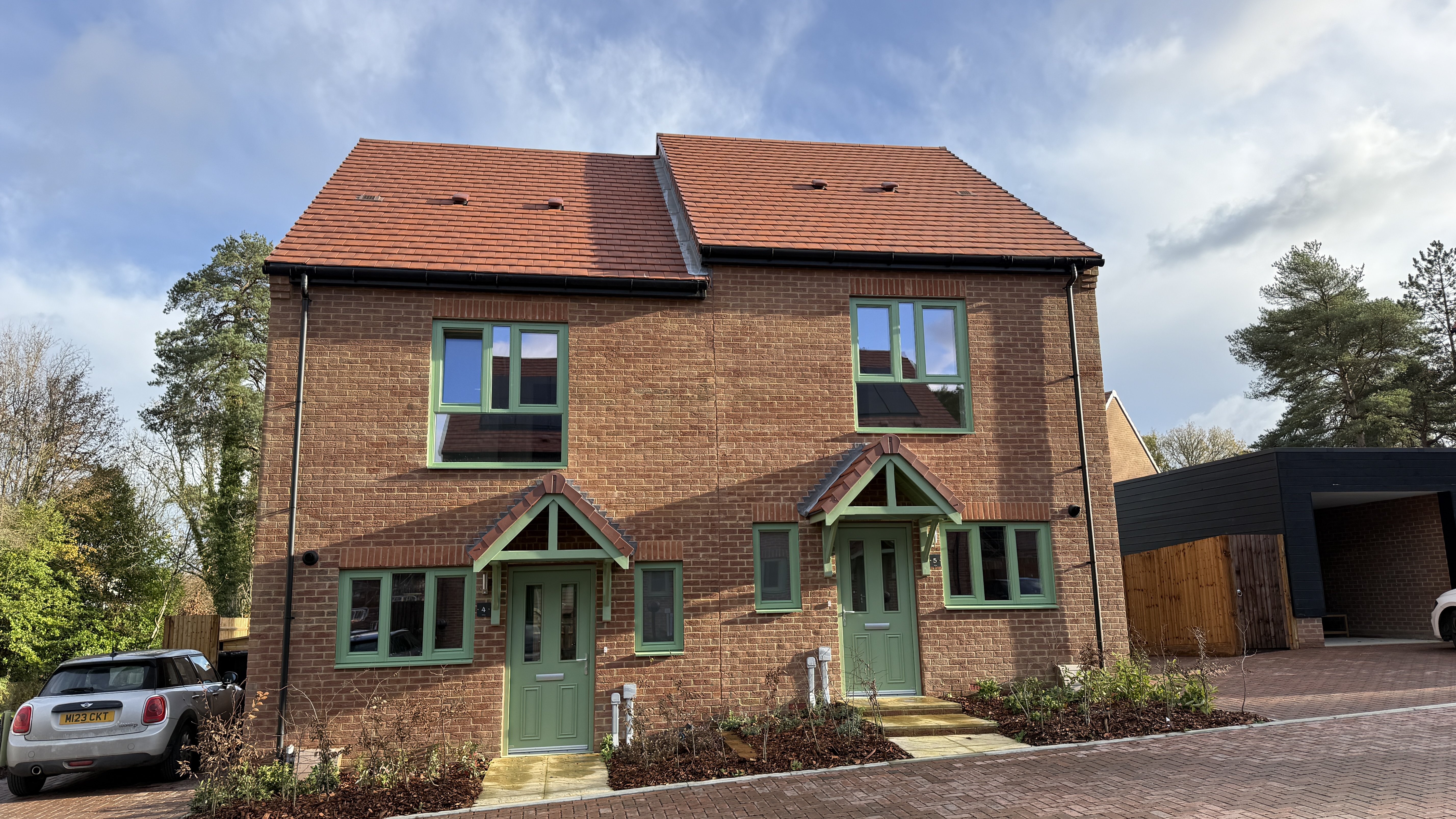 Front view of two modern brick houses with pitched tiled roofs, green front doors with small entrance canopies, white‑framed windows, and planted front gardens.