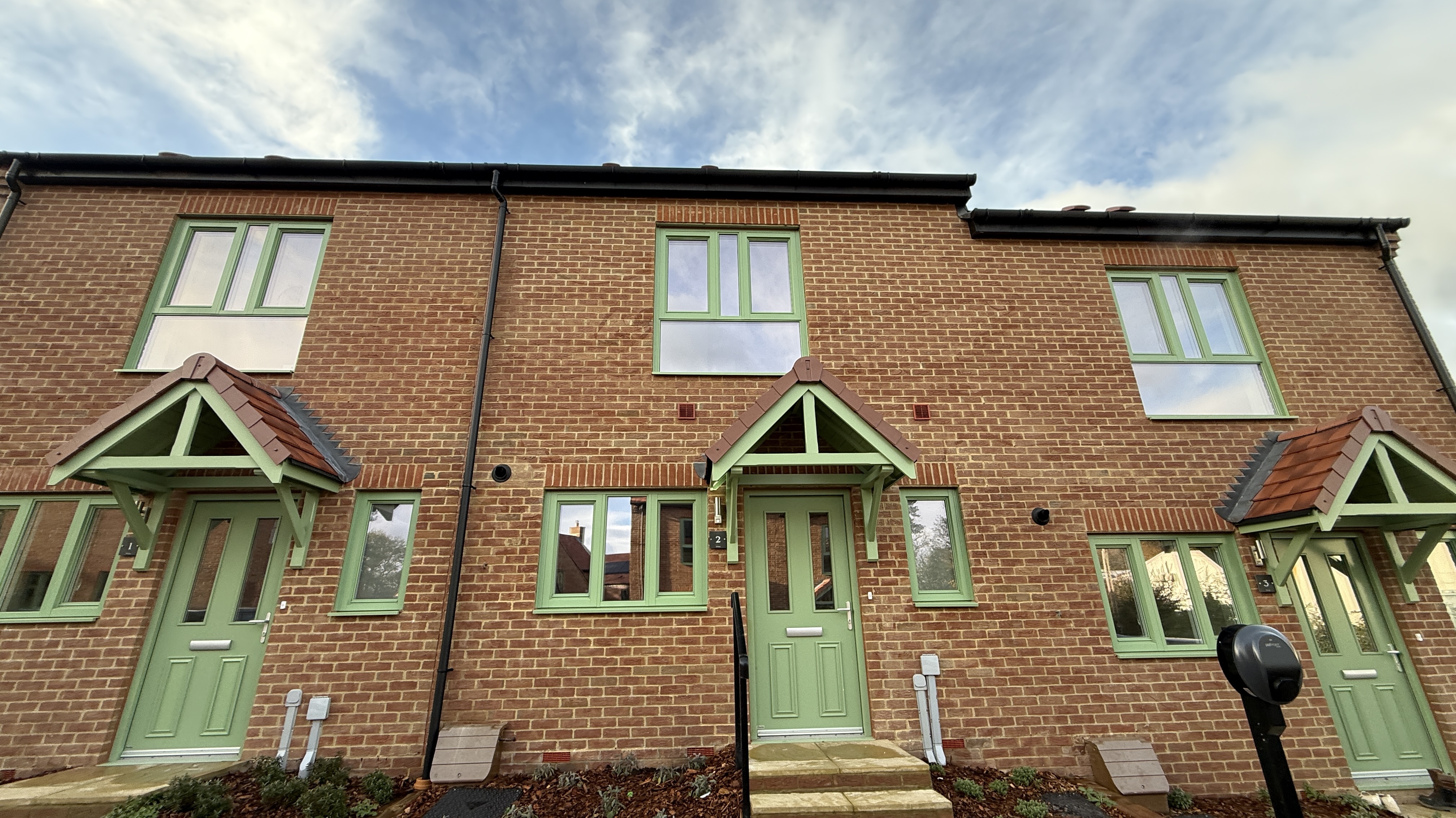 Front elevation of a modern brick house featuring green‑framed windows, a matching green front door with canopy, pitched roof, and a small planted frontage.