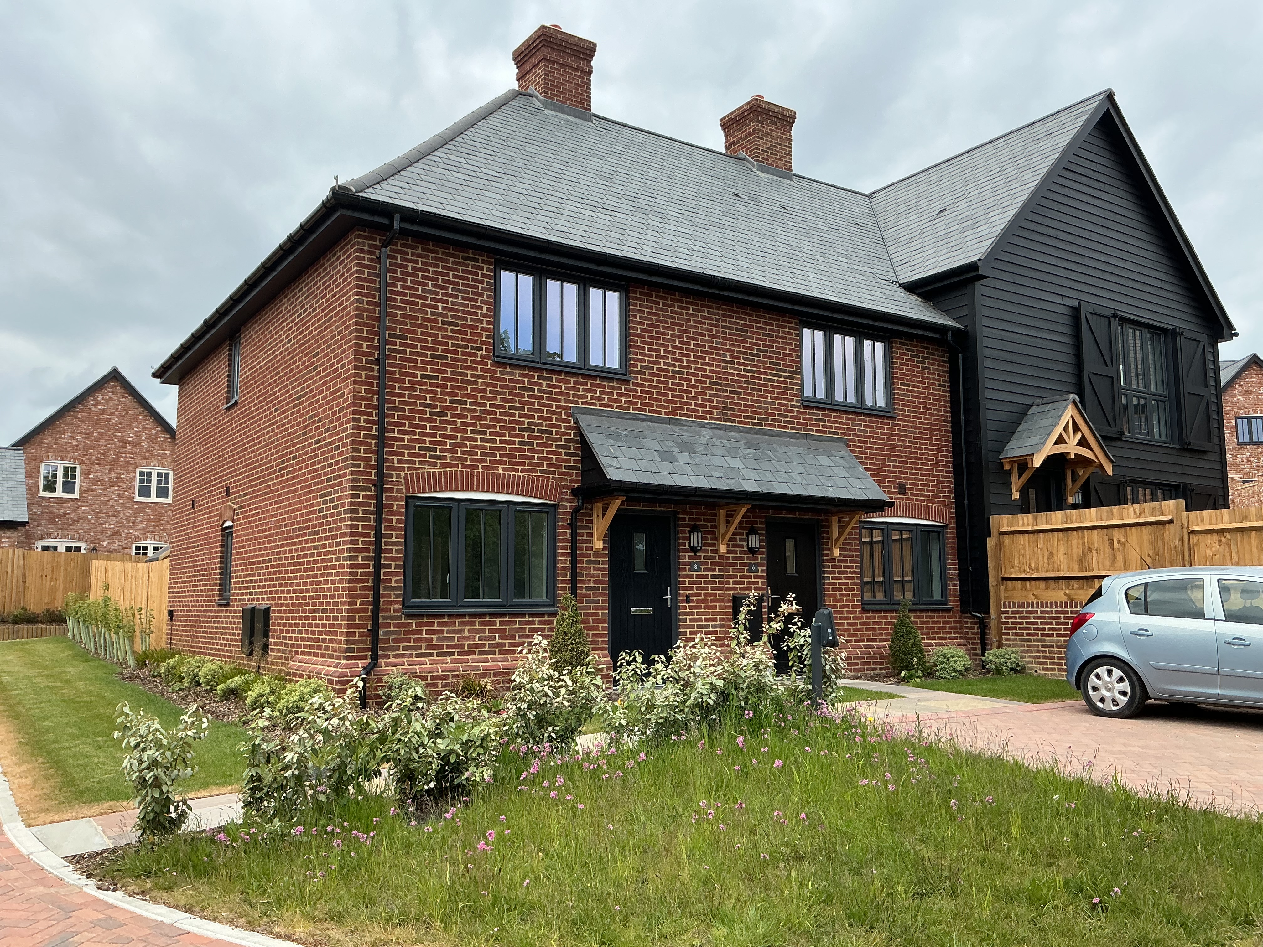 Modern red‑brick house with white‑framed windows, pitched roof, covered front entrance, and a small driveway area beside a planted front garden.