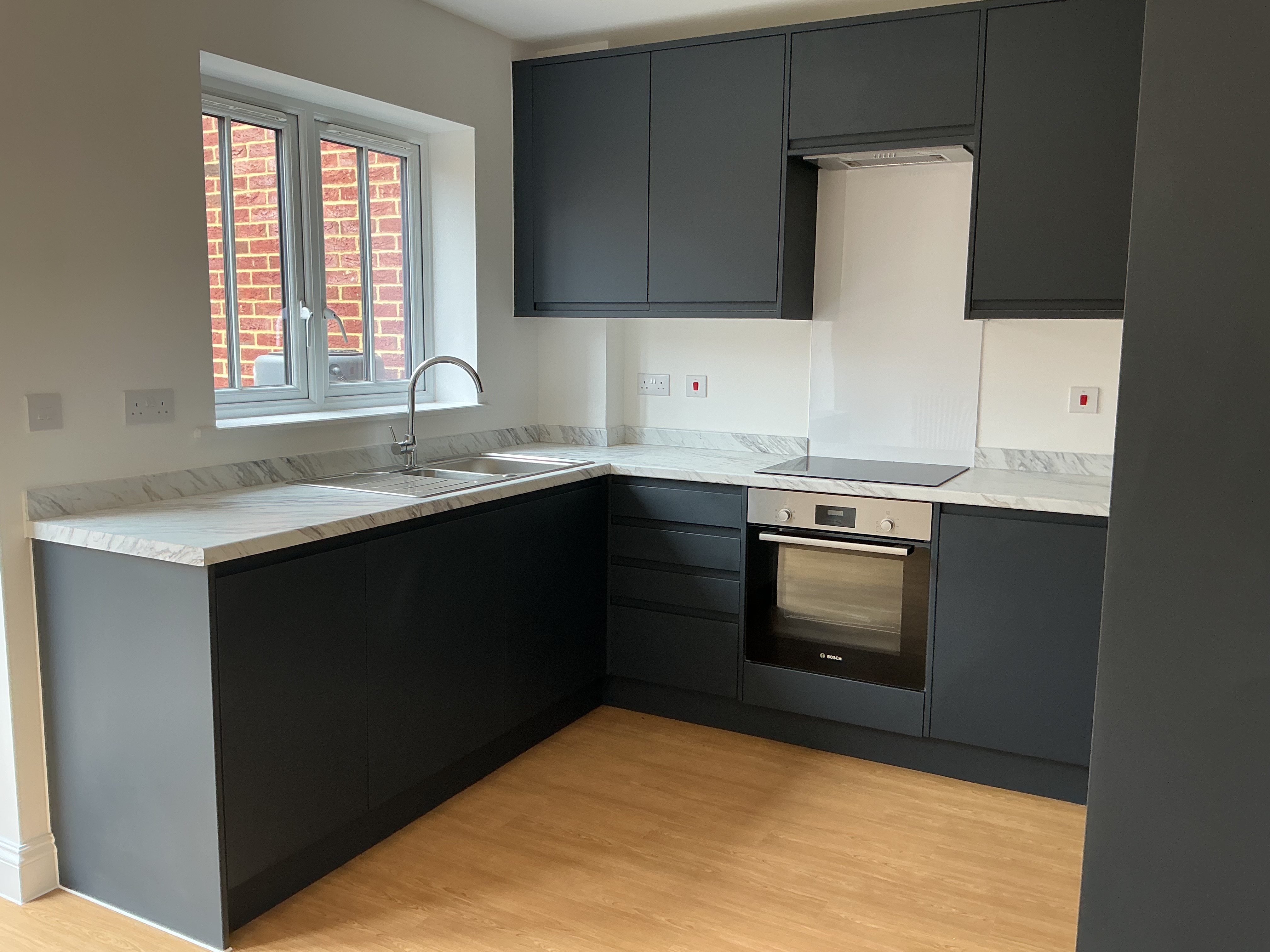 Modern fitted kitchen with dark matt cabinets, light worktops, integrated oven and hob, stainless steel sink, wood‑effect flooring, and a window providing natural light.