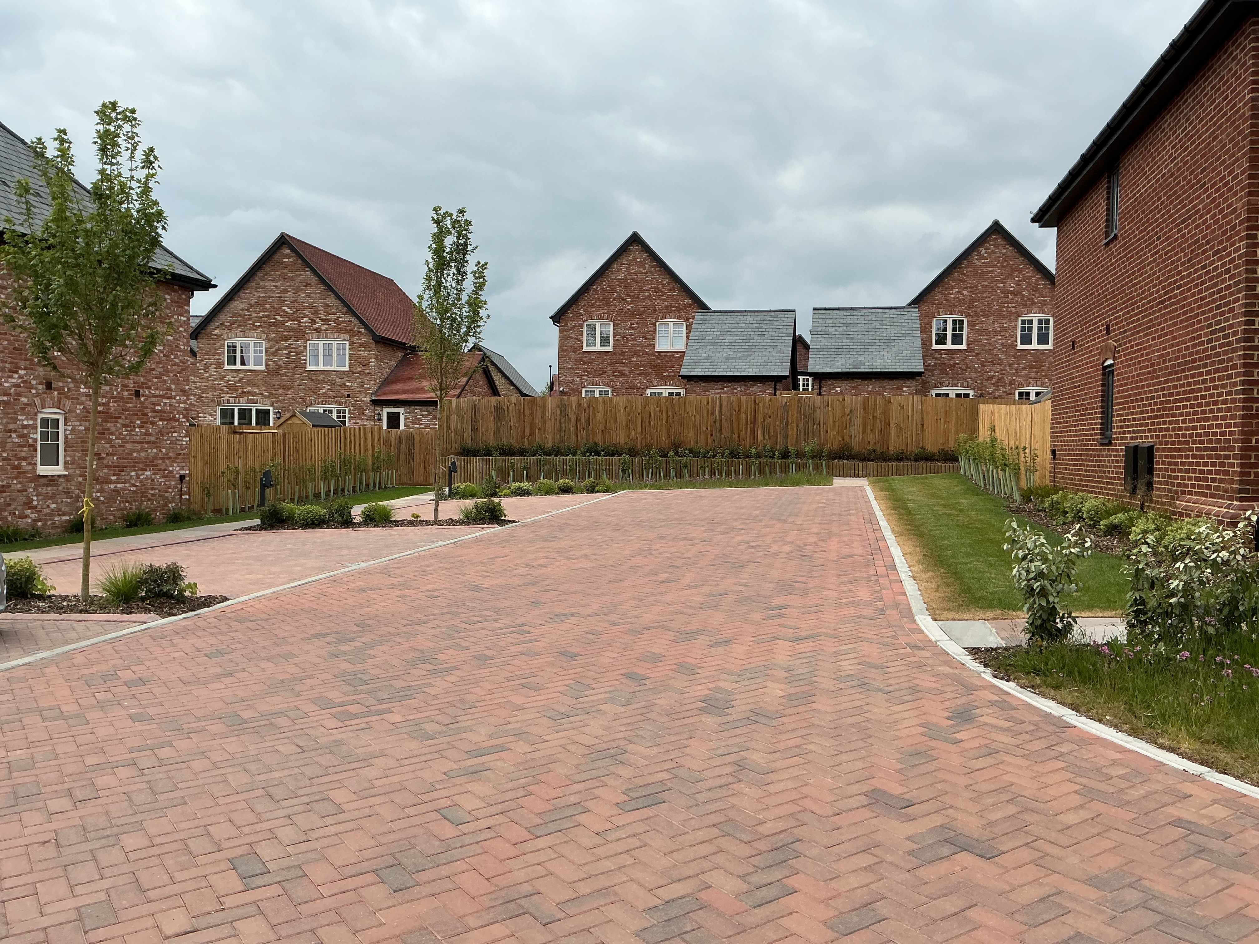 Brick‑paved residential courtyard with parking spaces, wooden boundary fencing, low planting, and modern brick houses with pitched roofs in the background.