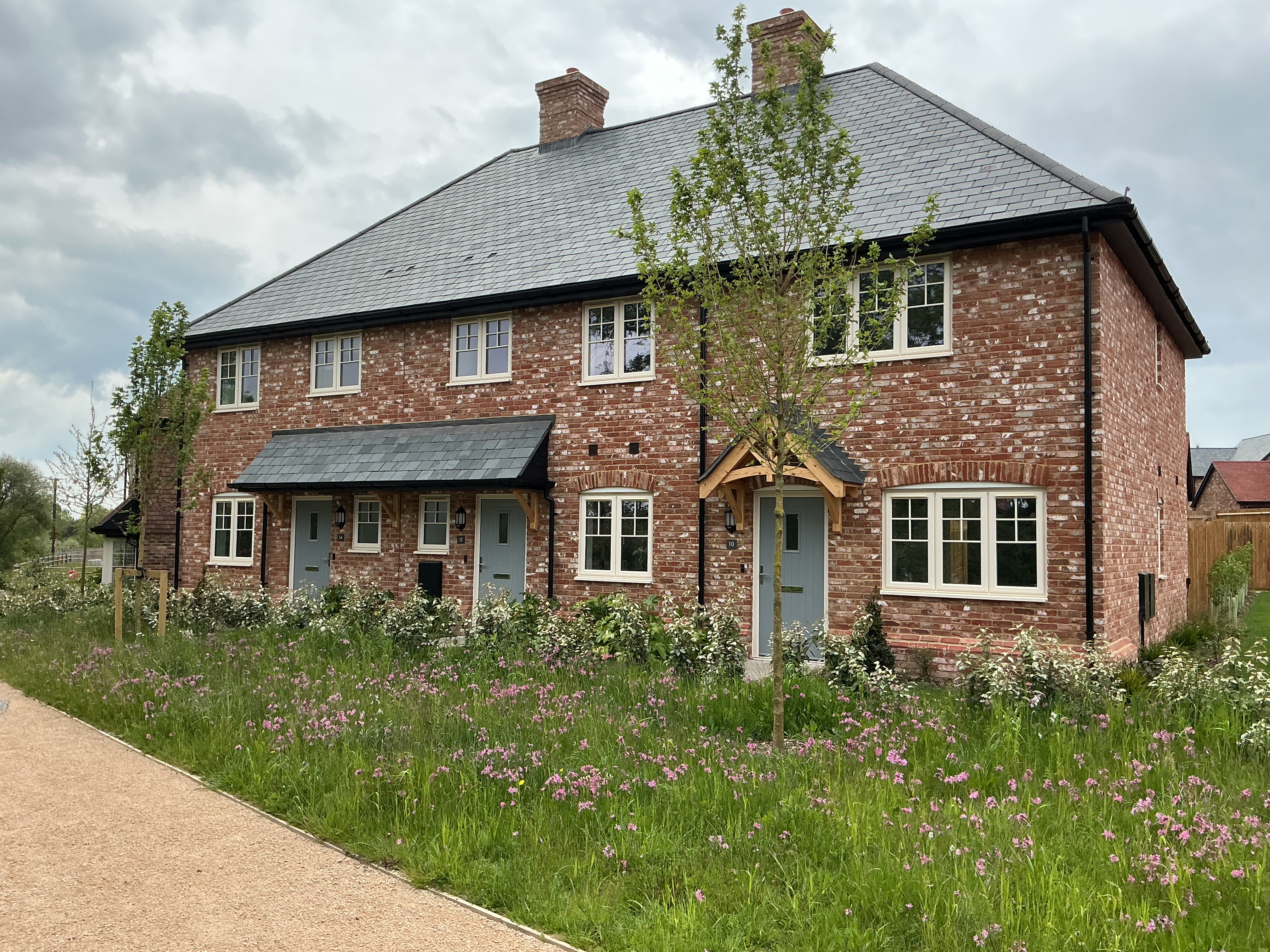 Front view of a modern red‑brick house with white‑framed windows, pitched tiled roof, small covered entrance, and a planted front garden with wildflowers and shrubs.