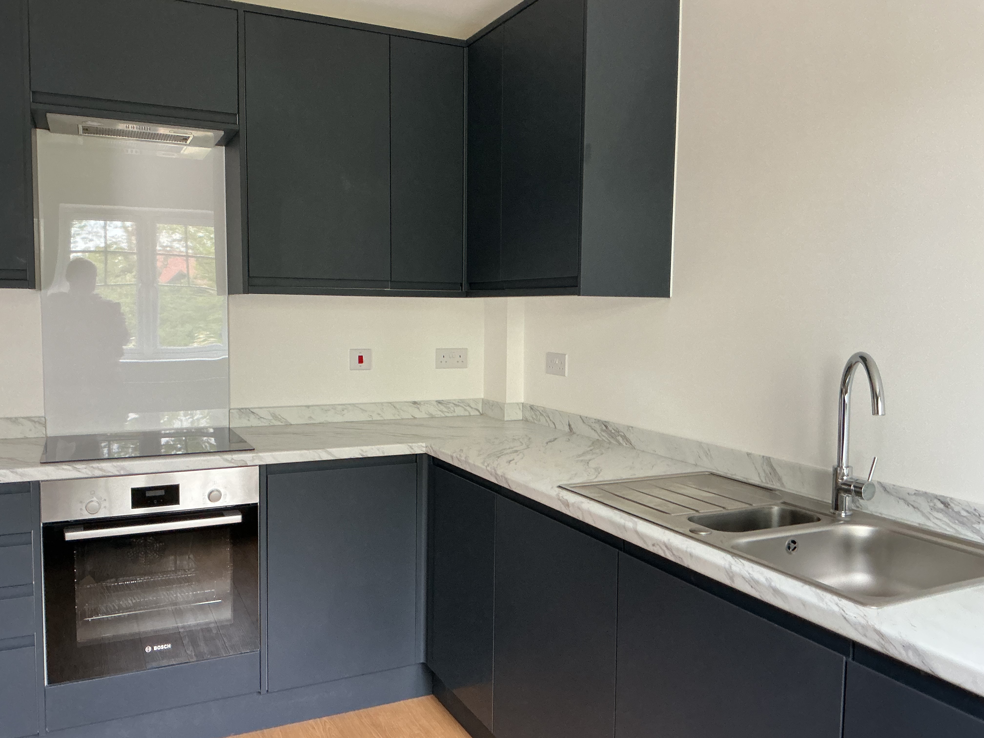Corner view of a modern kitchen featuring dark fitted cabinets, light worktops, built‑in oven, stainless steel sink, tiled splashback, and wood‑effect flooring.