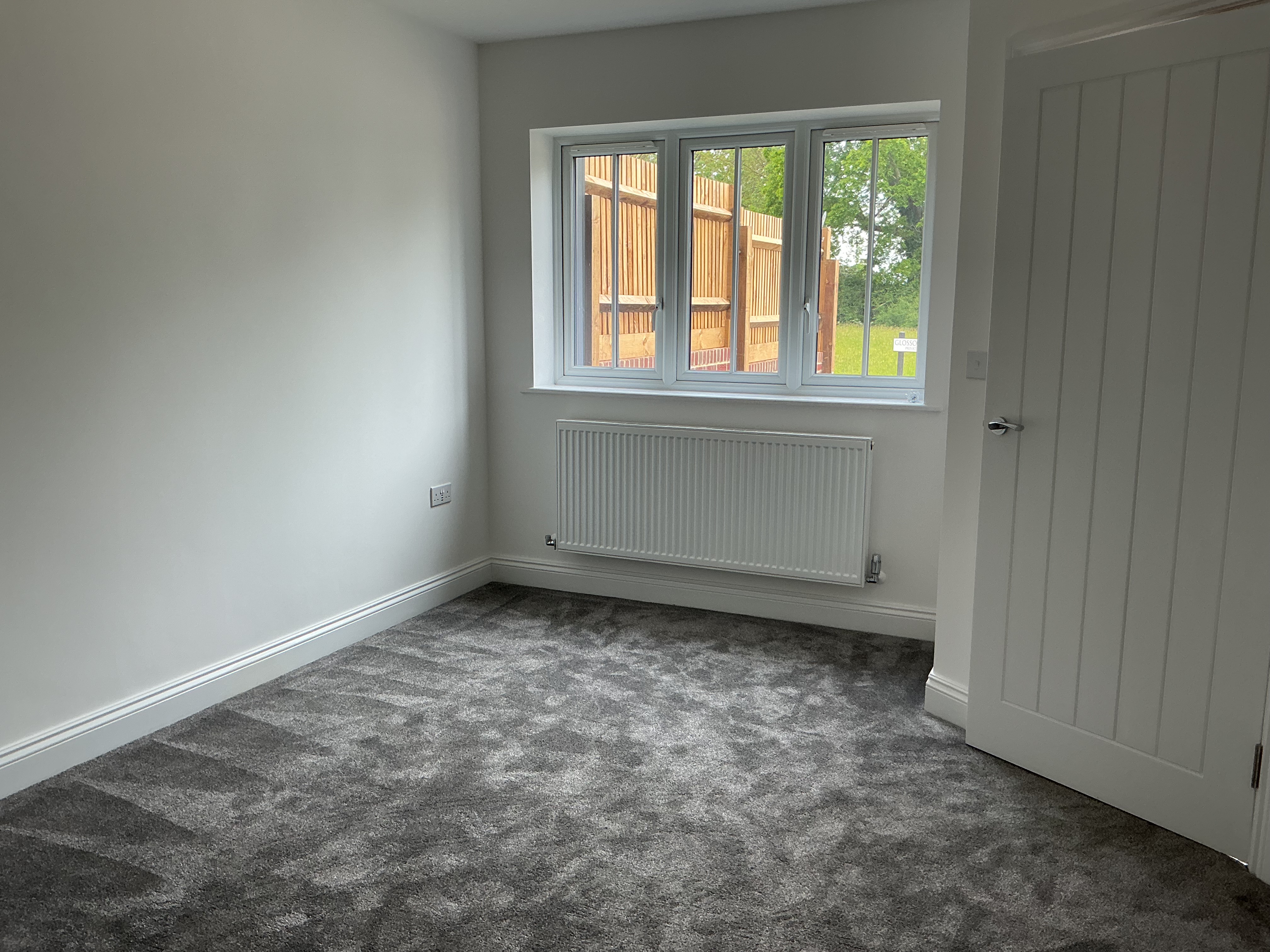 Bright, unfurnished room with grey carpeted flooring, neutral d&eacute;cor, a window with radiator below, and a white internal door.