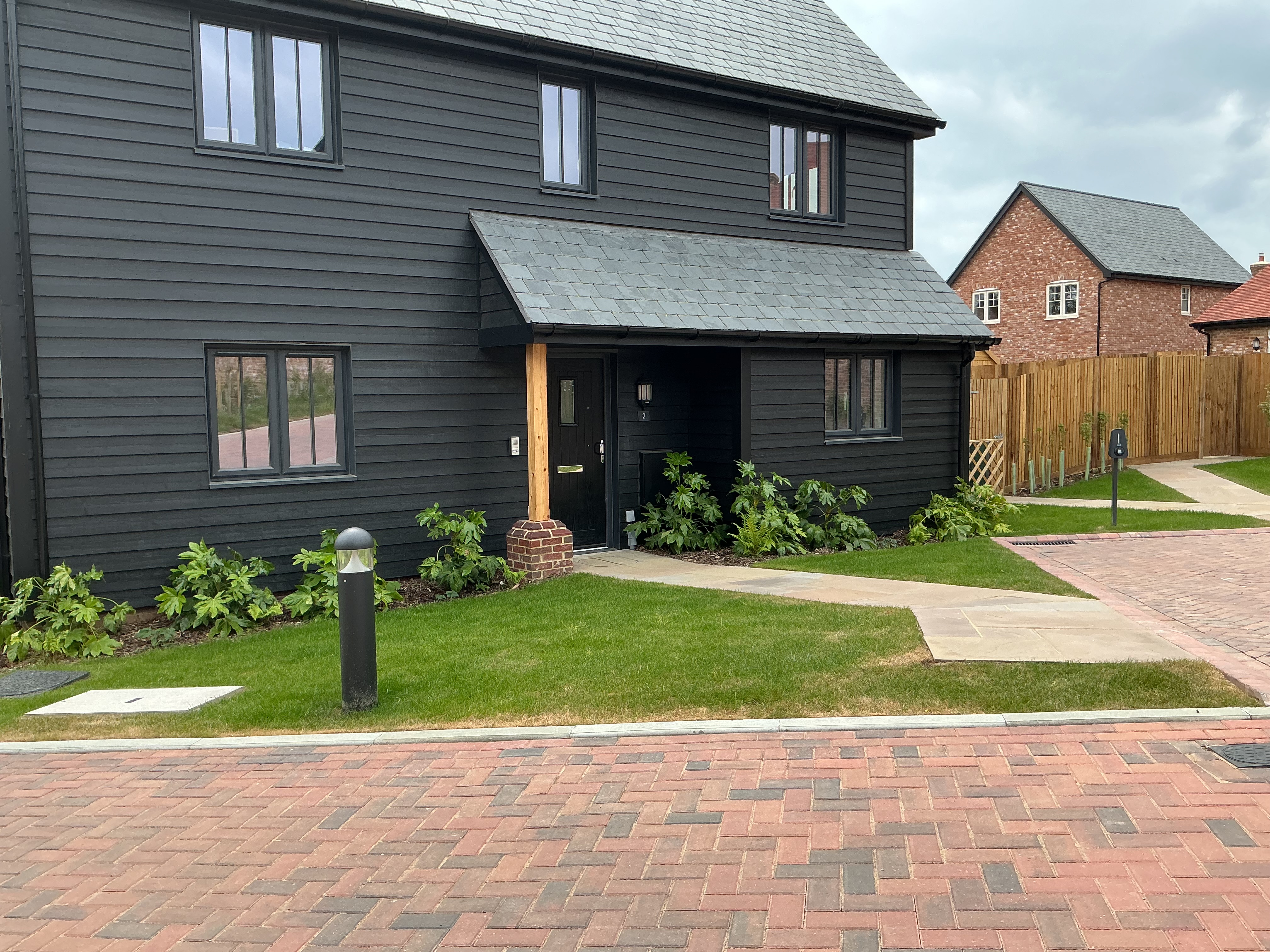 Contemporary house with dark external cladding, pitched roof, white‑framed windows, covered entrance, and a landscaped front garden with lawn and planted borders.