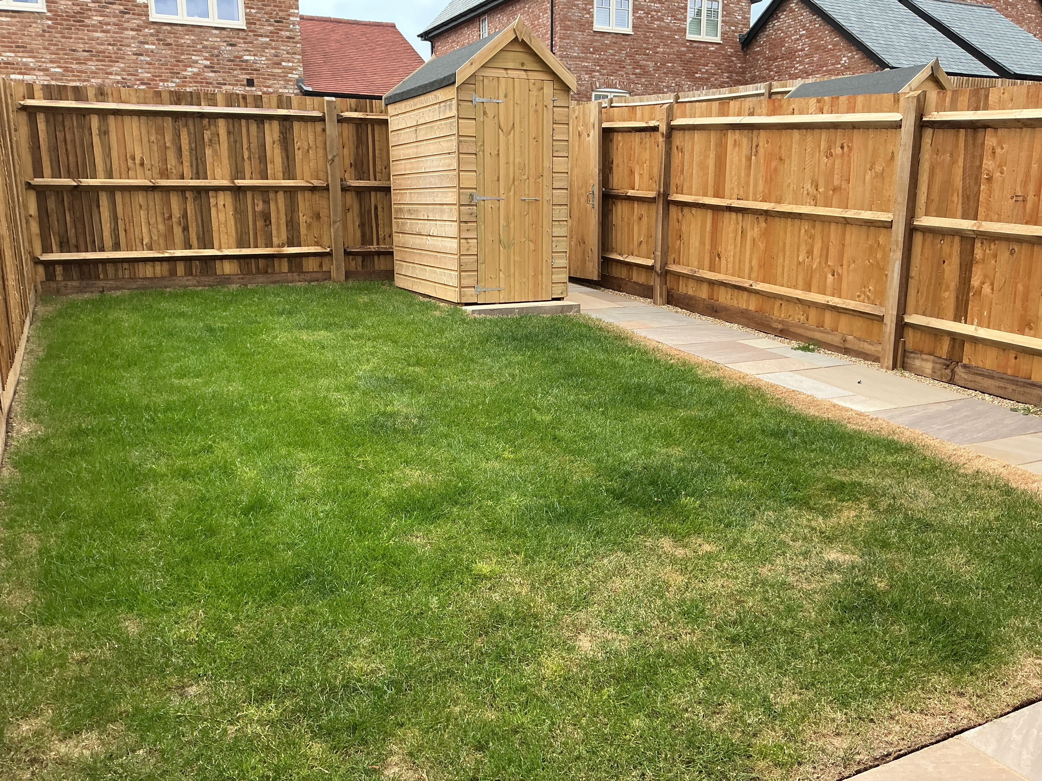 Private rear garden featuring a central grass lawn, wooden fencing, paved pathways along the edges, and a timber shed positioned at the far end.
