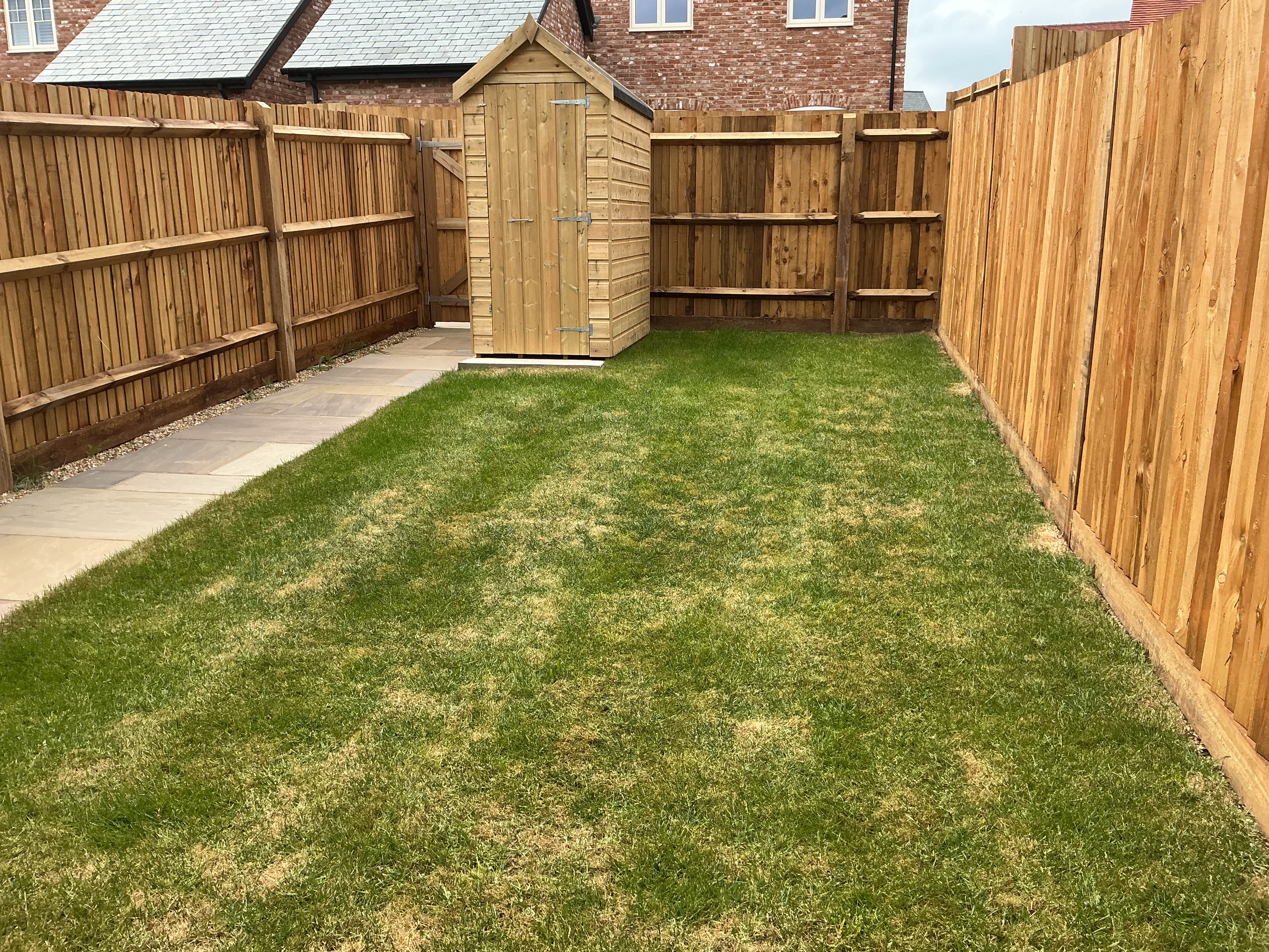 Enclosed rear garden with a grass lawn, timber fencing on all sides, a wooden garden shed at the rear, and paved side access alongside the lawn.