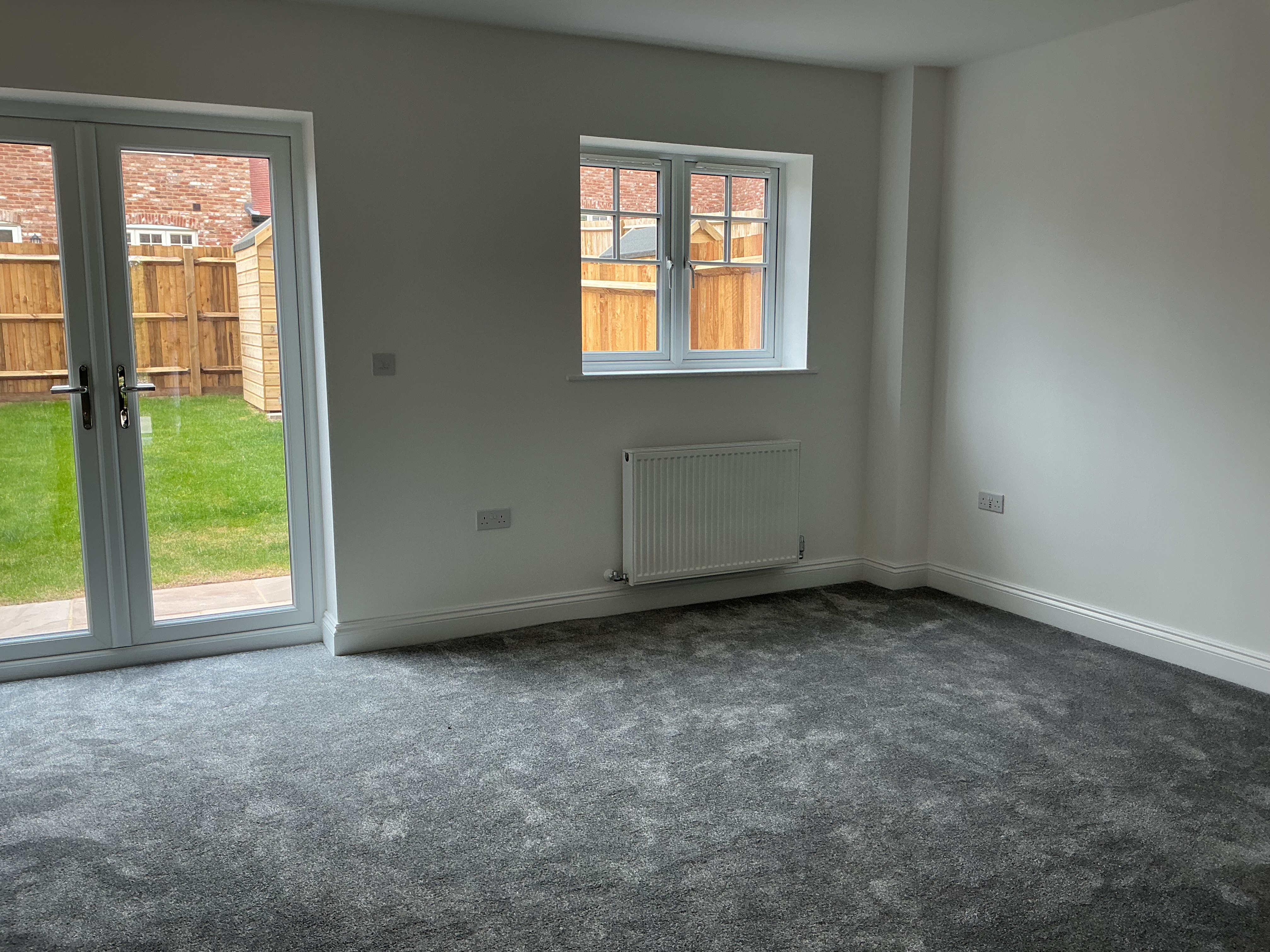Unfurnished living room with grey carpeted flooring, neutral walls, a window with radiator beneath, and glazed doors opening onto the rear garden.