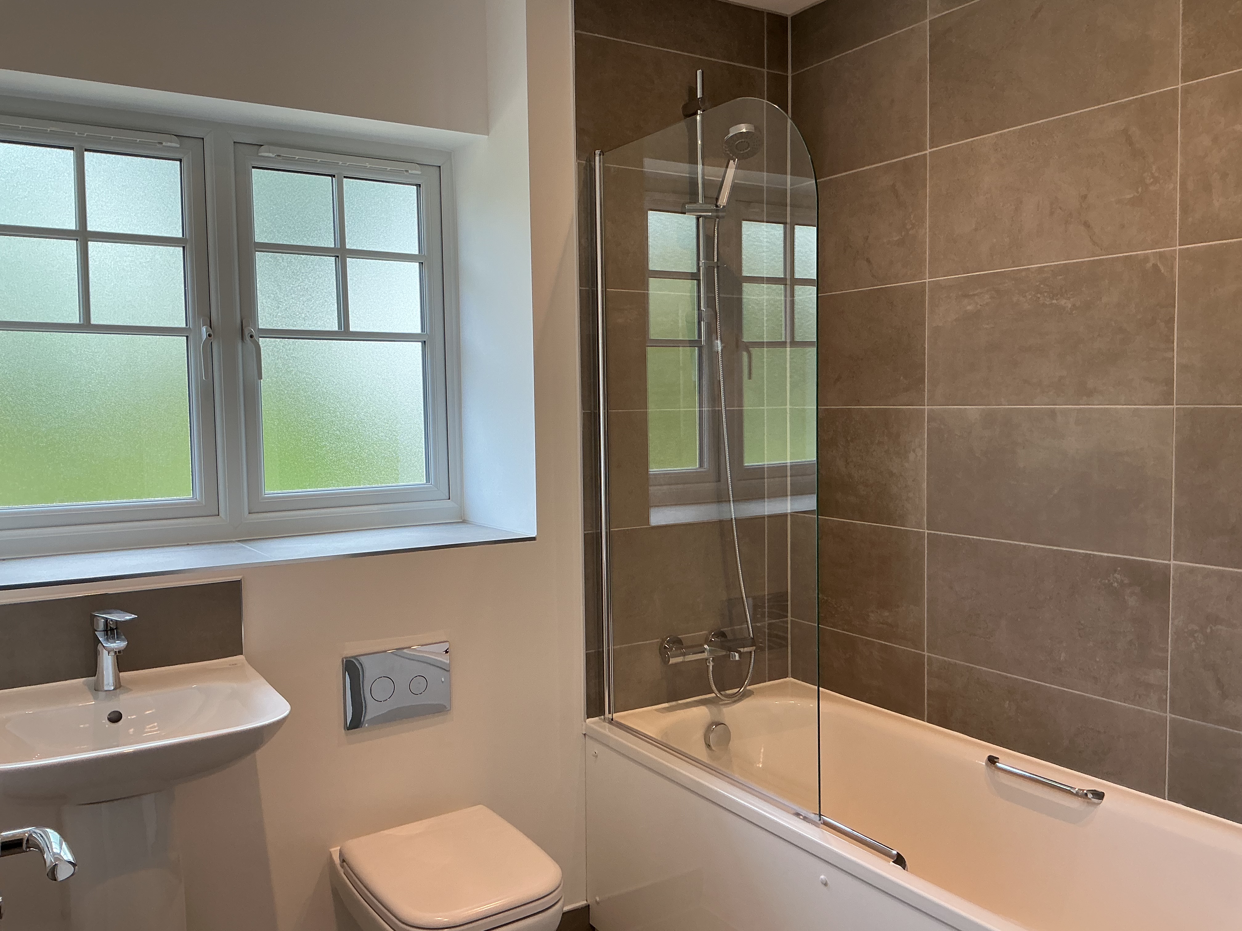 Modern bathroom with a full‑size bathtub and glass shower screen, tiled walls, toilet, wall‑mounted flush plate, wood‑effect flooring, and a frosted window providing natural light.