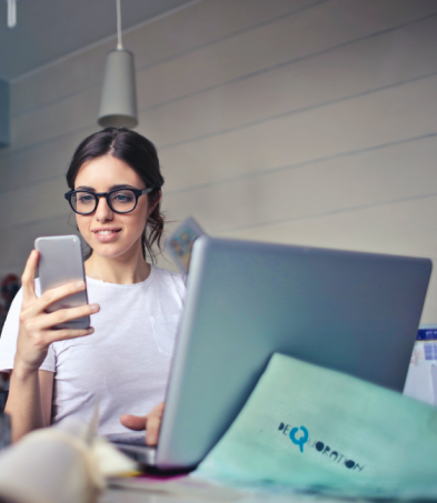 Woman using a smartphone while working on a laptop at a desk.