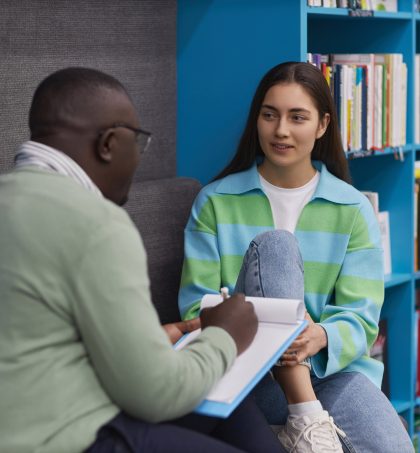 A tutor sits facing a learner with a notebook in hand as they talk in a library or study area, with bookshelves in the background.