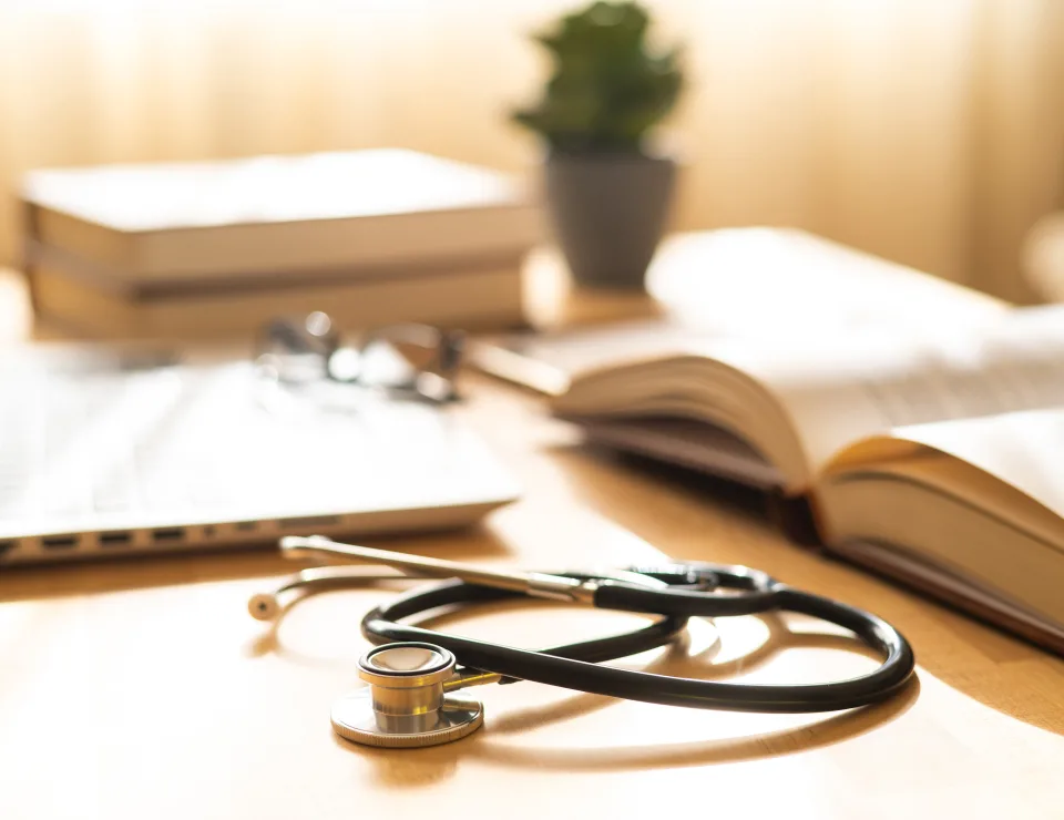 Stethoscope resting on a wooden desk next to an open book, laptop, and blurred eyeglasses, symbolizing medical education, healthcare study, and professional development in a warm, inviting setting