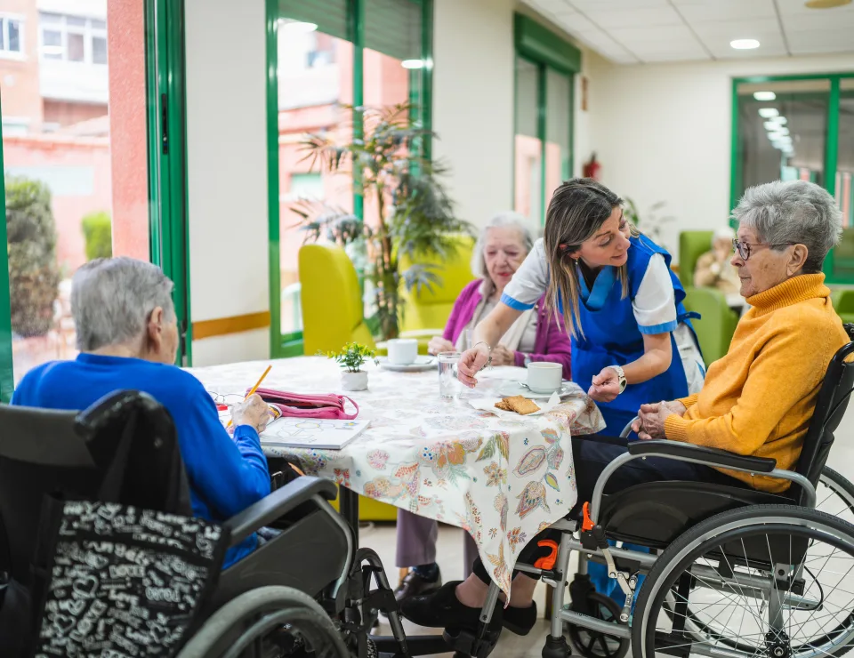 Caregiver assisting three senior women in wheelchairs at a table during an activity in a bright, modern nursing home setting