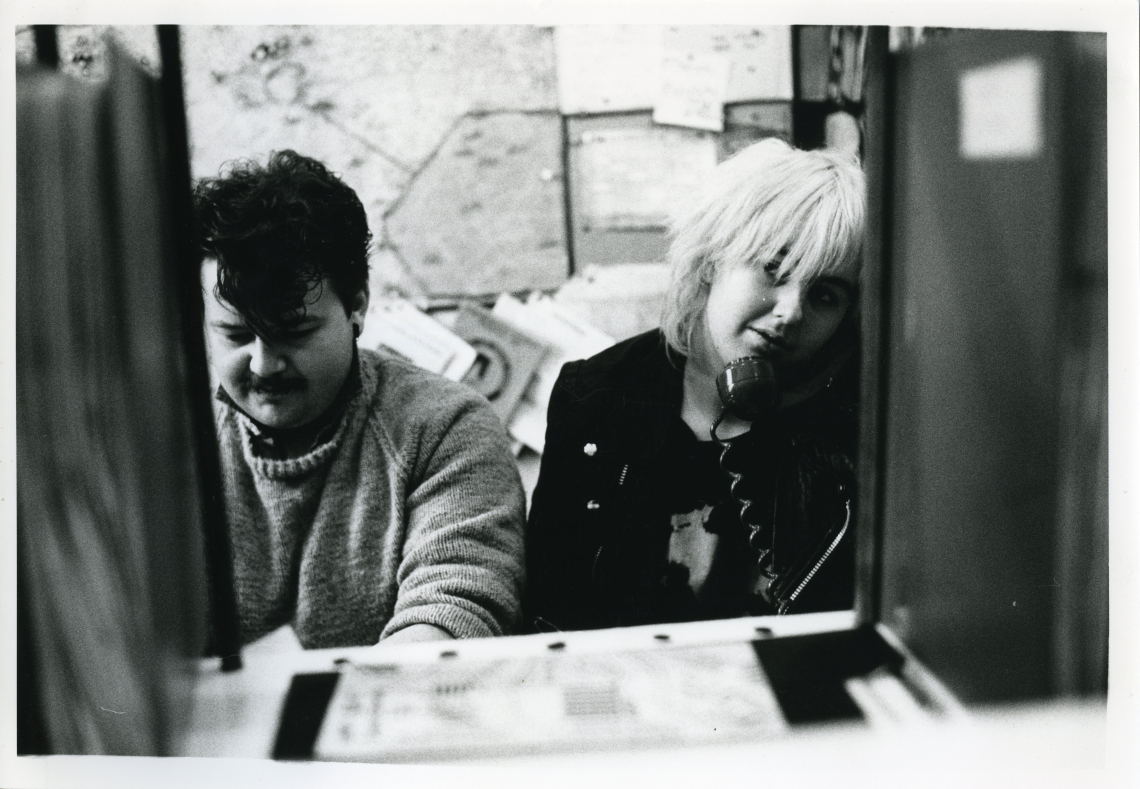 Two people in the Switchboard office, who are making calls. A noticeboard is behind them.