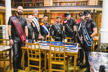A photo from the opening of the UK Leather & Fetish Archive at Bishopsgate Institute. Eight men are standing round a table in the Library, dressed up in leather.