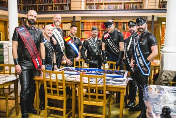 A photo from the opening of the UK Leather and Fetish Archive at Bishopsgate Institute. Eight men are standing round a table in the Library, all dressed in leather.