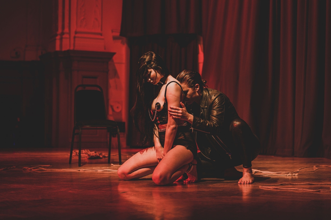 Two people engage in the aftercare section of a bondage session. The woman in the front who has been bound is on her knees in fetish gear