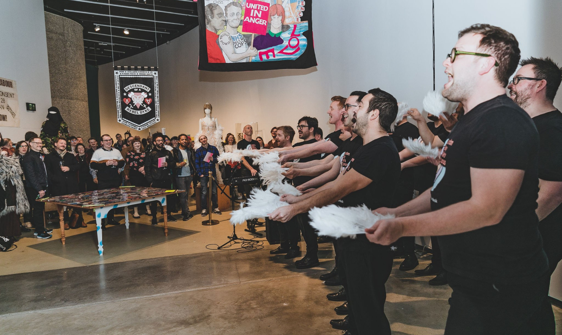 A photograph of members of London's Gay Men's Chorus performing at the exhibition opening. they each are holding a feathered fan in each hand and are using them for emphasis whilst singing in front of a large audience.