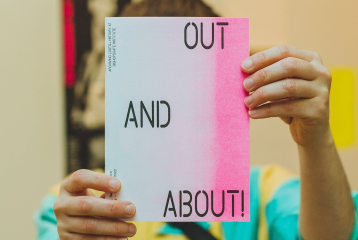 A person holding over their face a zine from the Out and About! exhibition