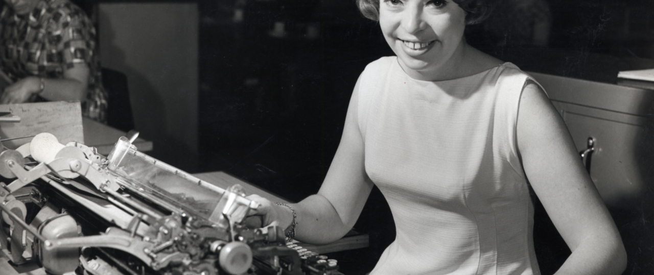 A black and white image of a woman sat smiling by a typewriter.