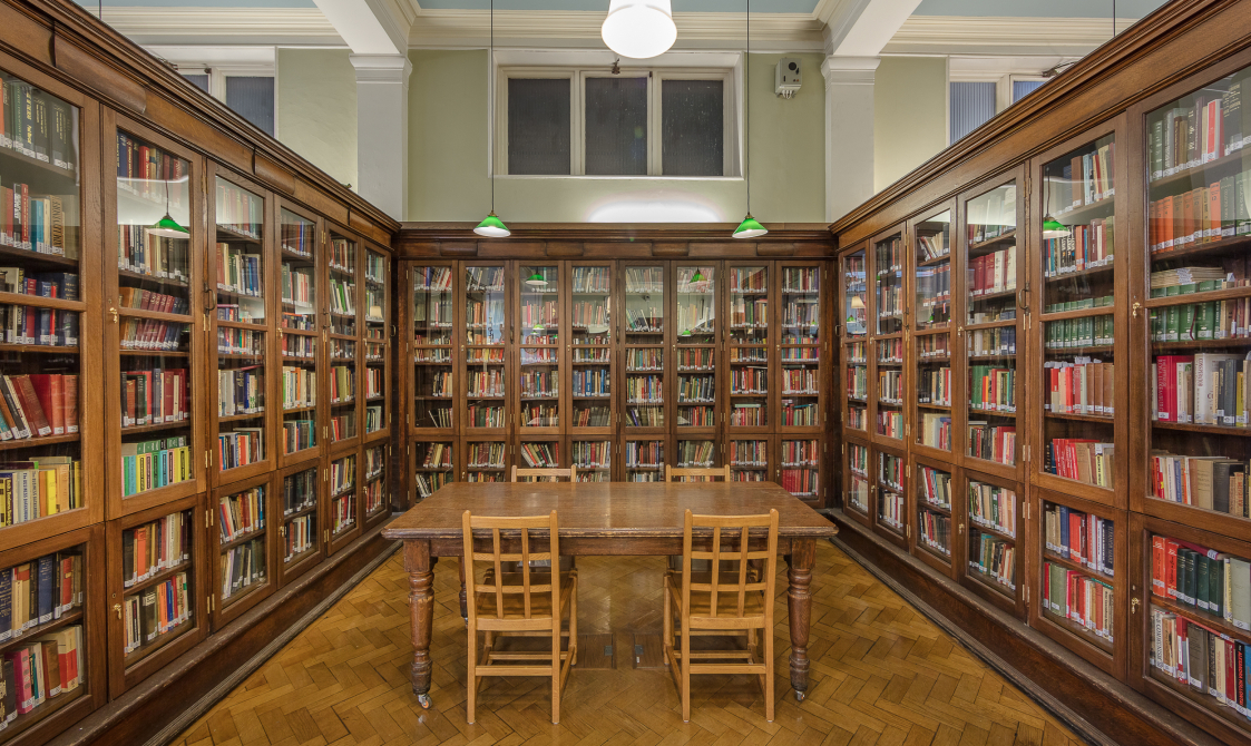 A photo of Bishopsgate Institute Library. Three cabinets with books are in the left hand side, right hand side, and the back of the photo. A wooden desk is in the middle of these cabinets, with four chairs around the table.