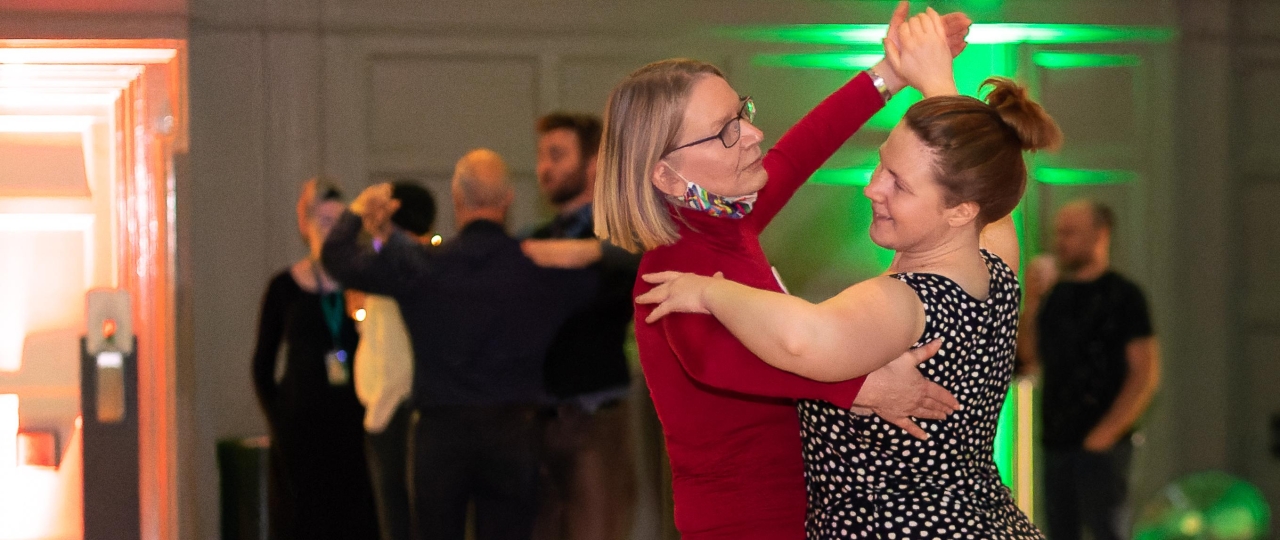 Two women dance together in the Great Hall of Bishopsgate Institute. In the background other same-sex couples can be seen dancing.