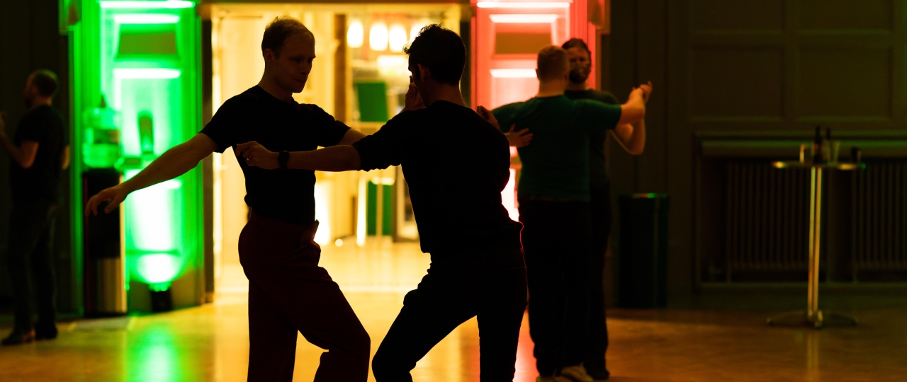 Two men engage in a striking dance pose in the Great Hall at Bishopsgate Institute. We can only see their silhouettes, as they are standing in front of green and red coloured background lighting.