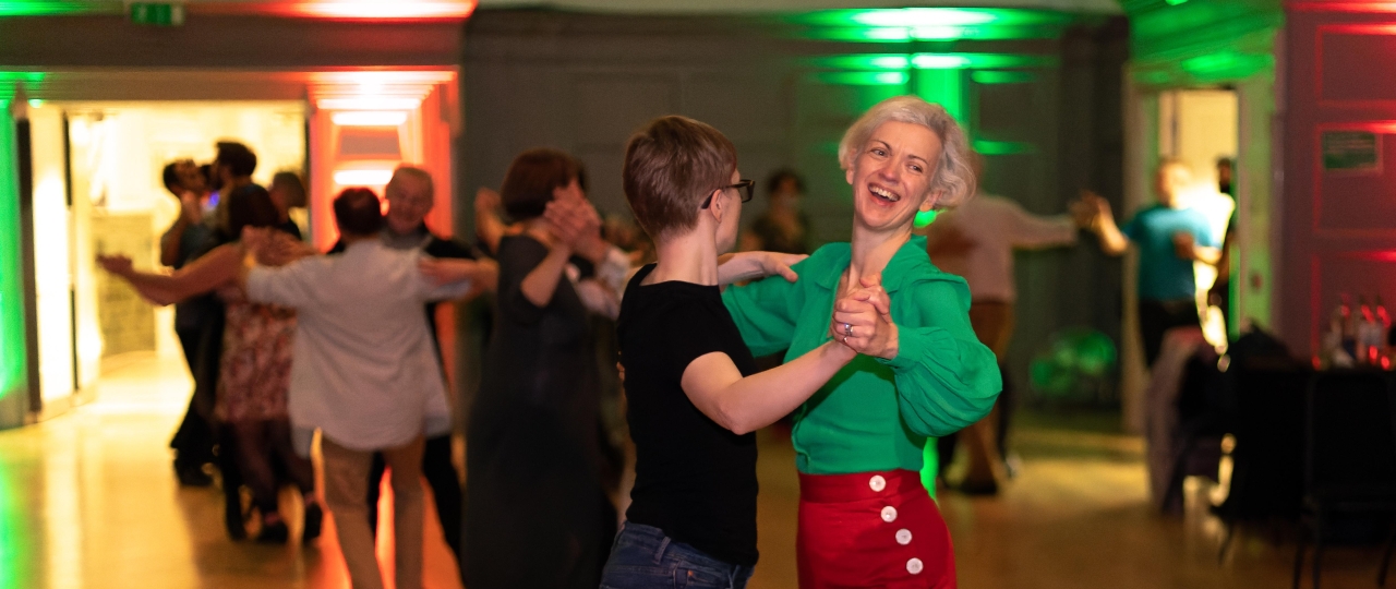 Two women engage in a dance pose in the Great Hall at Bishopsgate Institute. The woman on the left is dressed in black and is turned away from the camera, whilst the blonde woman on the right is smiling and wearing colourful clothing. In the background, other same-sex couples can be seen dancing.