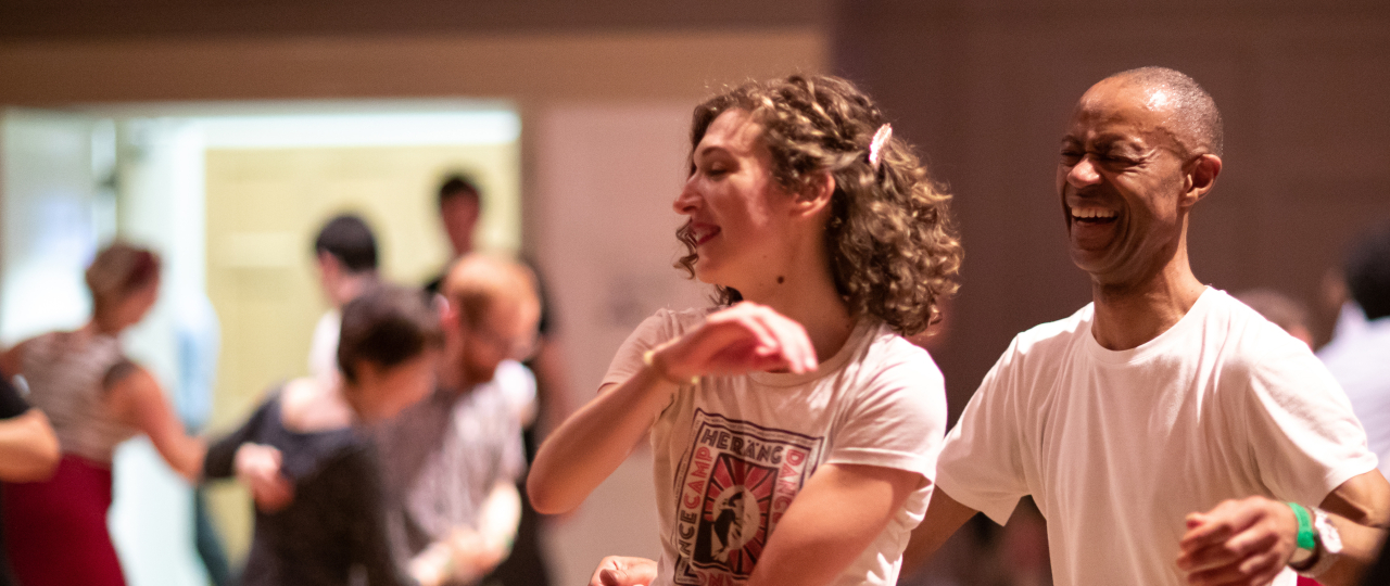 Two Swing Dancers, one man on the right, and one woman on the left, are laughing and dancing together. In the background, out of focus, other Swing Dancers can be seen in the Great Hall at Bishopsgate Institute.