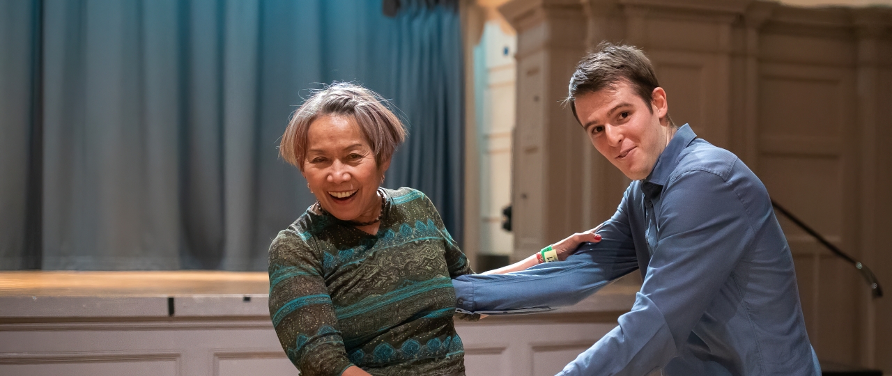 Two Swing Dancers in front of the stage in the Great Hall at Bishopsgate Institute. The woman on the left is holding the hands of the younger man on the right.