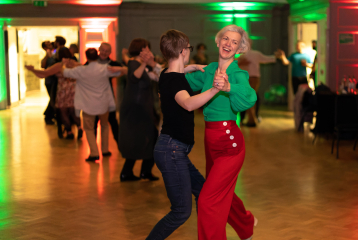 Two women engage in a dance pose in the Great Hall at Bishopsgate Institute. The woman on the left is dressed in black and is turned away from the camera, whilst the blonde woman on the right is smiling and wearing colourful clothing. In the background, other same-sex couples can be seen dancing.