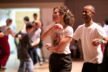 Two Swing Dancers, one man on the right, and one woman on the left, are laughing and dancing together. In the background, out of focus, other Swing Dancers can be seen in the Great Hall at Bishopsgate Institute.