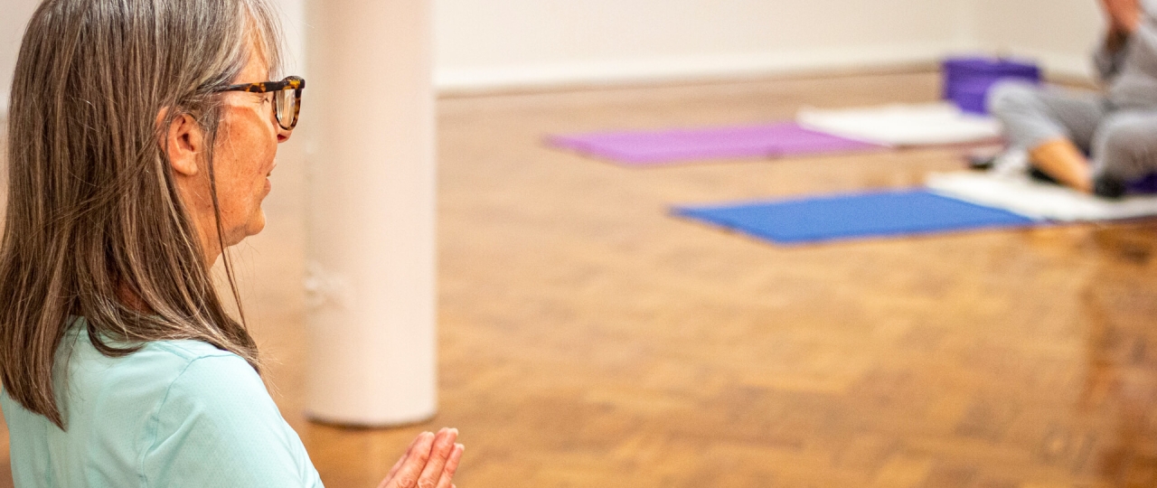 A photo from a Iyengar yoga session at Bishopsgate Institute. The teacher is at the front of the room, with the students opposite her. They are sitting on yoga mats with their legs crossed and palms together.