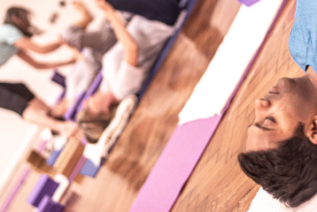 A photo from an Iyengar yoga workshop at Bishopsgate Institute. Students are lying down on their mats, and the teacher is going round, adjusting their posture.
