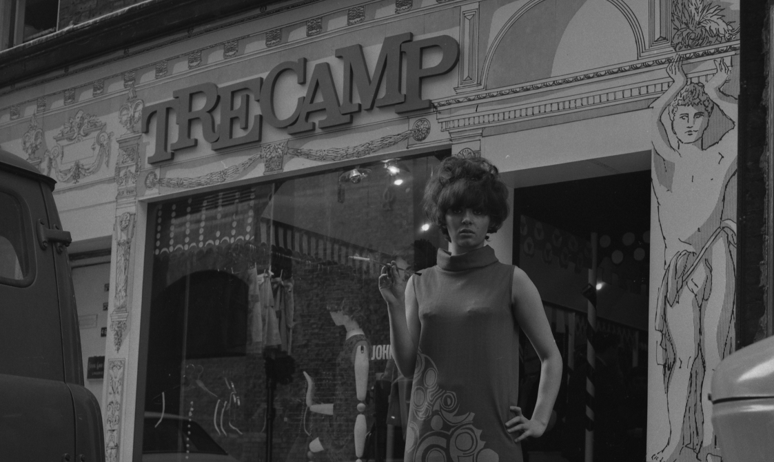 A woman is standing outside the shop Tree Camp in the 1960s. She is wearing a long dress and has her left shoulder on her left hip and is holding a cigarette holder in her right hand.