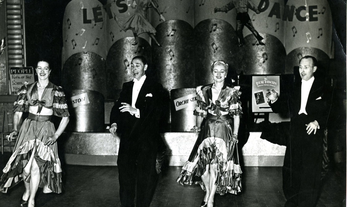 Two women and two men dancing, with the women in flamingo style dresses. Behind them is a sign that says: "Let's dance".
