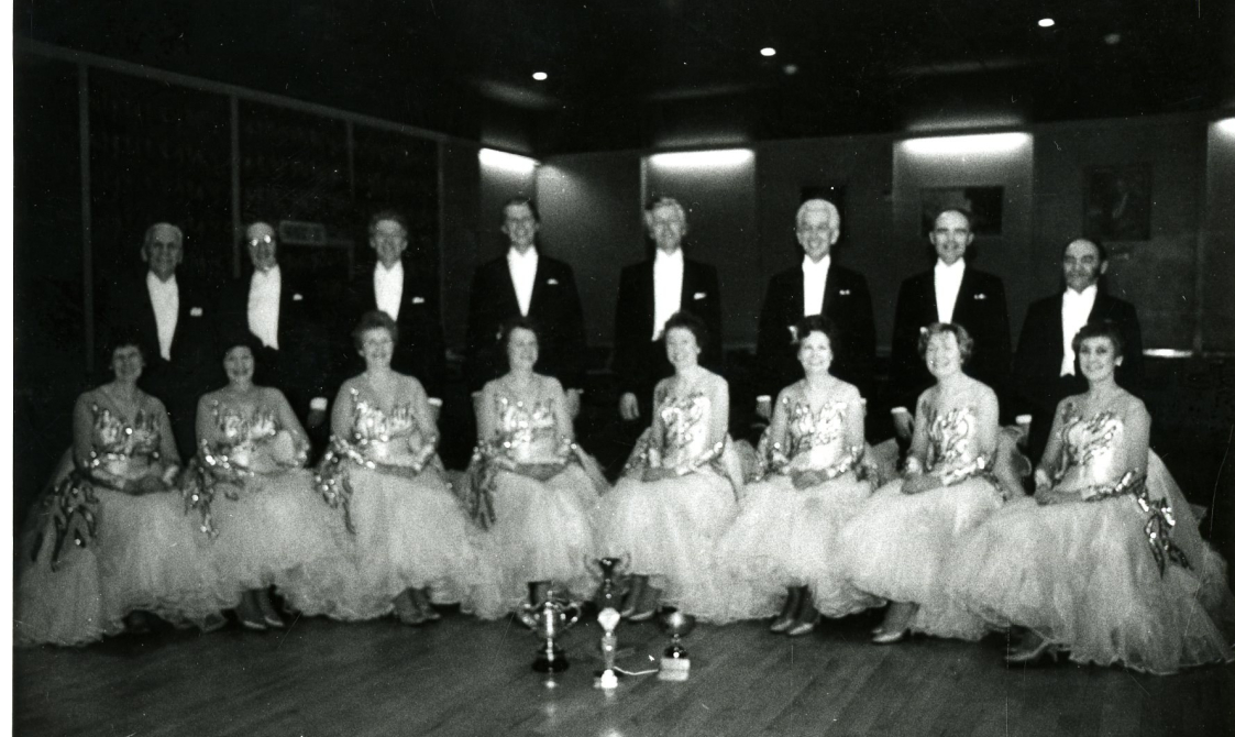 Nine dancing pairs kneeling on a ballroom floor, with the trophy in front of them.