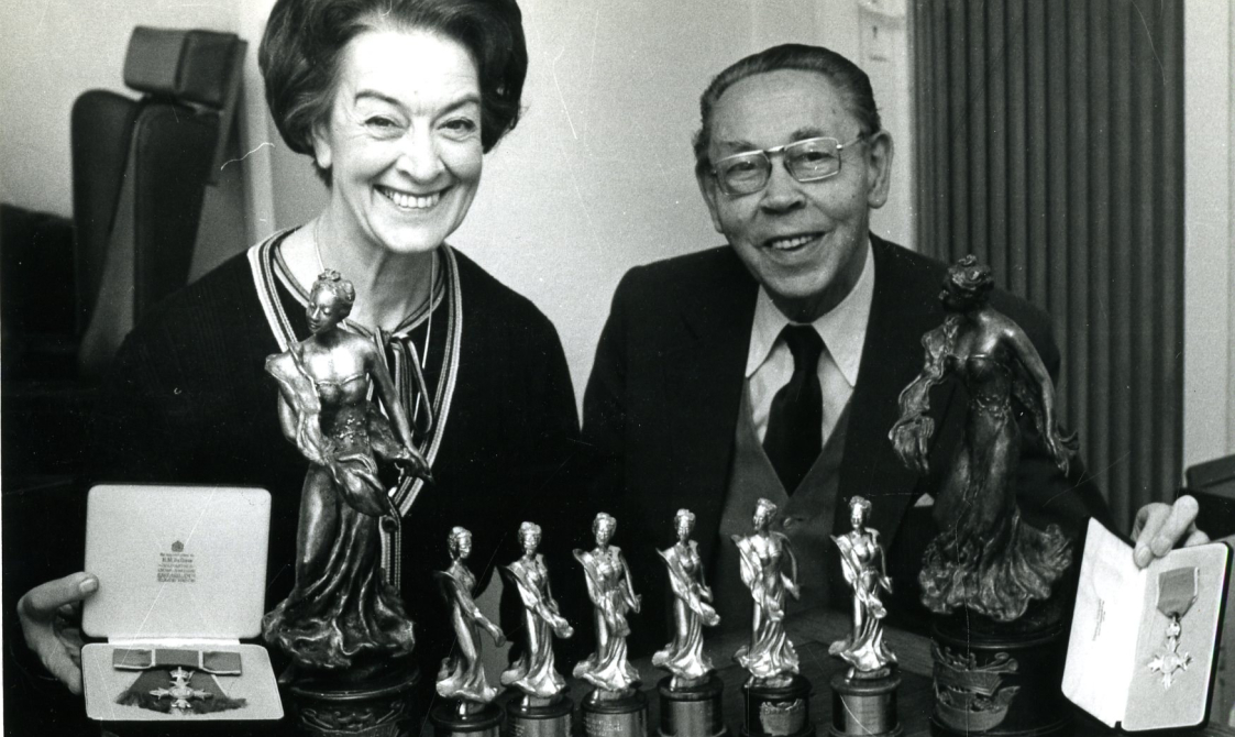 Peggy Spencer and her husband Frank sitting down with various trophies in front of them.