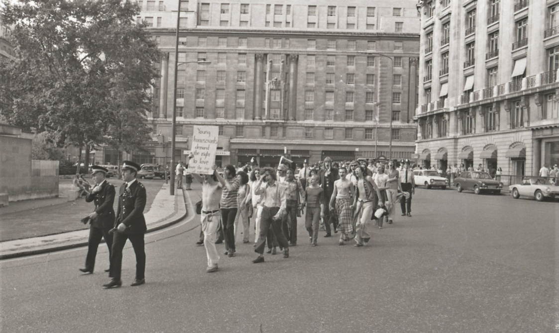 A picture from the second Pride march in 1973. Marchers are walking down the road, with police in front. The people at the front are holding a sign that says: "Young homosexuals have the right to love".