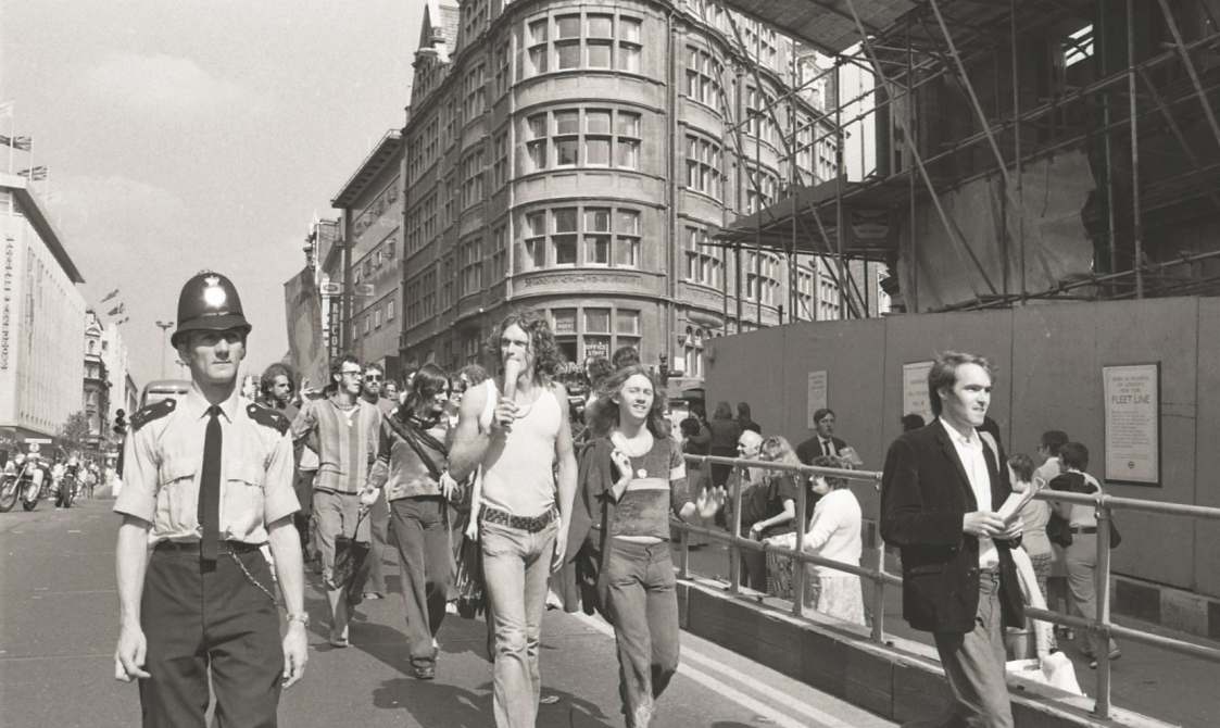 A picture from the second Pride march in 1973. Marchers are walking down the road in Central London, with a police officer next to them.