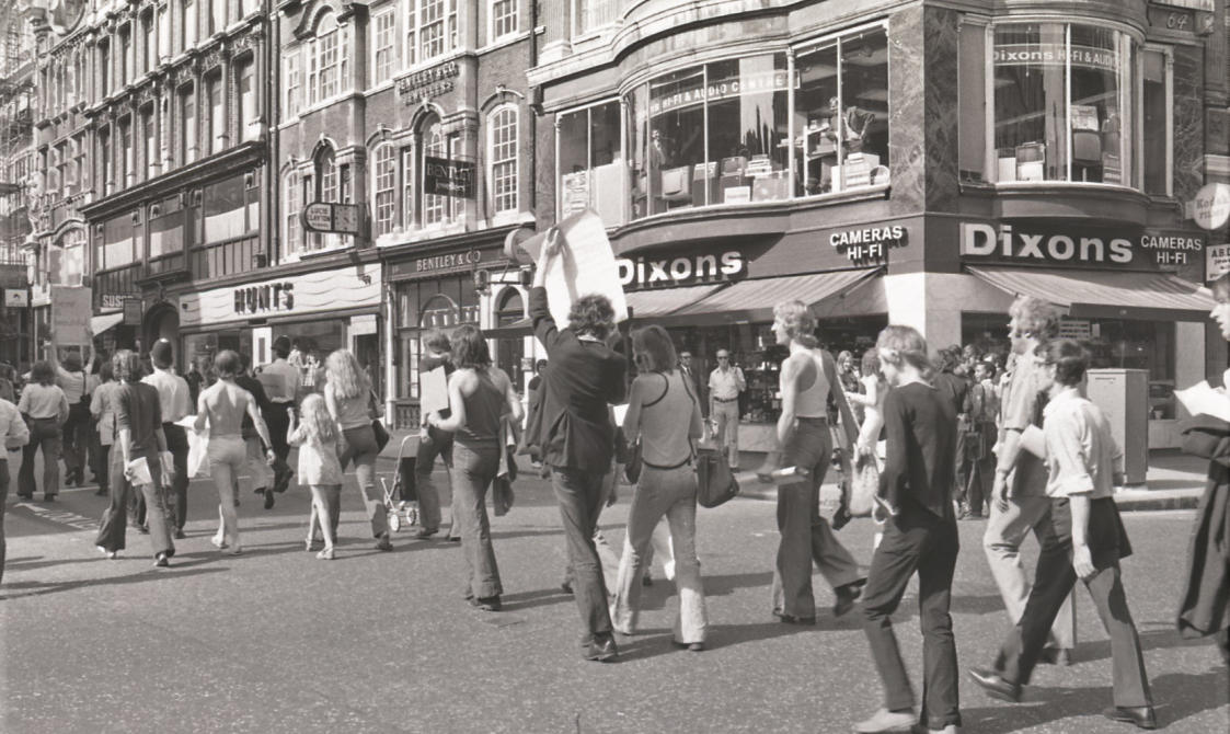 A picture from the second Pride march in 1973. Marchers are walking down the road in Central London, going past the shop Dixons.
