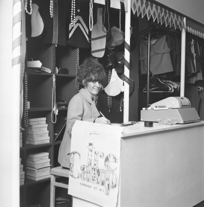 A woman working on the till at Tree Camp in the 1960s. Clothing railings are behind her and a till is on the desk in front of her.
