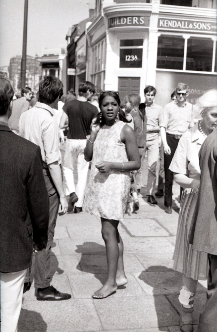 A black and white photo of King's Road in June 1966. A woman is standing in the middle of a group of people, facing the camera. She is wearing a shift dress and flat shoes.