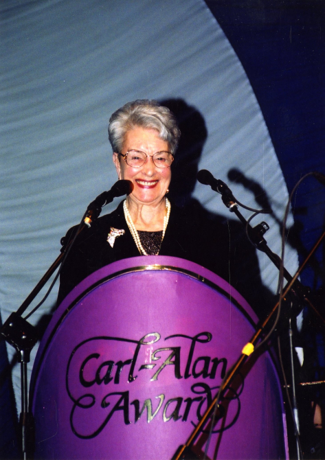 A picture of Peggy Spencer making a speech on stage. On the podium, it says: "Carl Alan Awards".