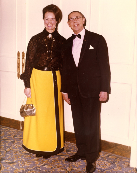 Peggy Spencer and her husband Frank. Frank is wearing a suit and Peggy is to his right, wearing a black shirt and yellow skirt.