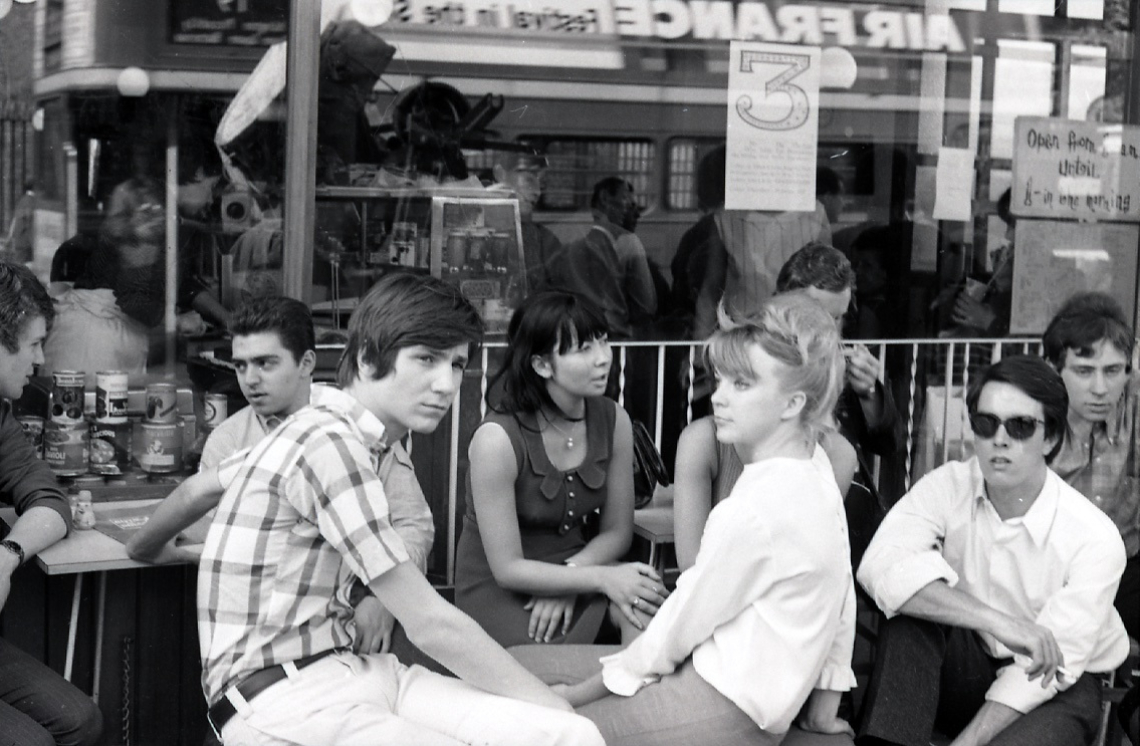 A group of young people are sitting outside a cafe. The image is black and white and was taken in the 1960s by Derek Brook.